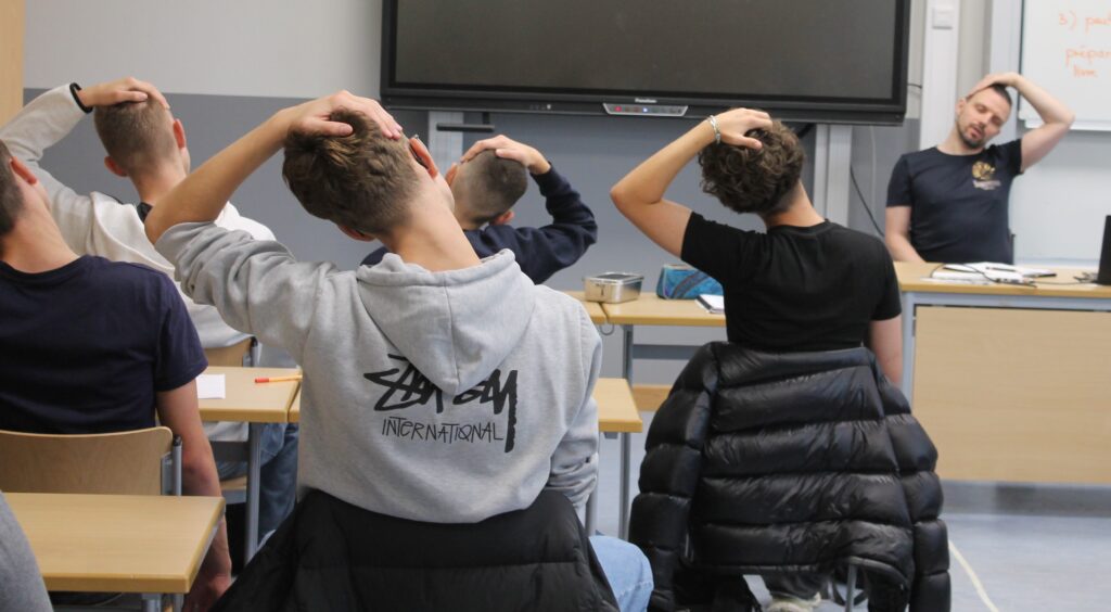 Das Bild zeigt einige sitzende Schüler von hinten, die an ihren Plätzen eine Yoga Übung ausführen.
